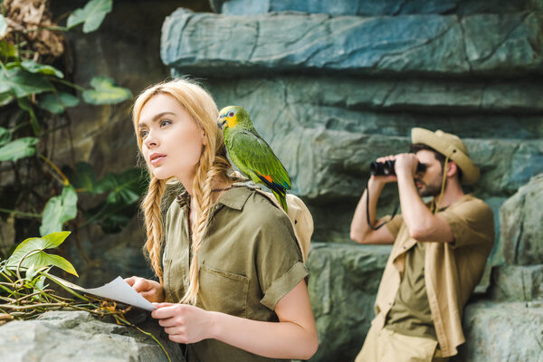 beautiful young woman in safari suit with parrot and map navigating in jungle while her boyfriend looking through binoculars