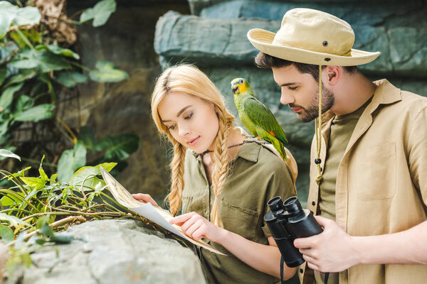 привлекательная молодая пара в сафари-костюмах с попугаем, пытающимся ориентироваться в джунглях
