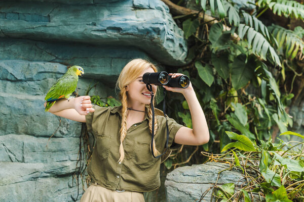 happy young woman in safari suit with parrot perching on arm looking through binoculars in jungle