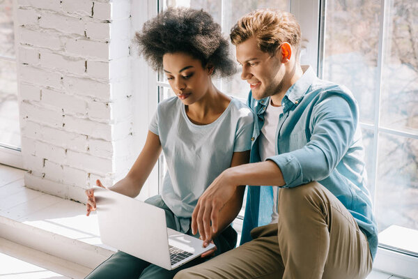 Multiracial couple looking at laptop screen while sitting by window