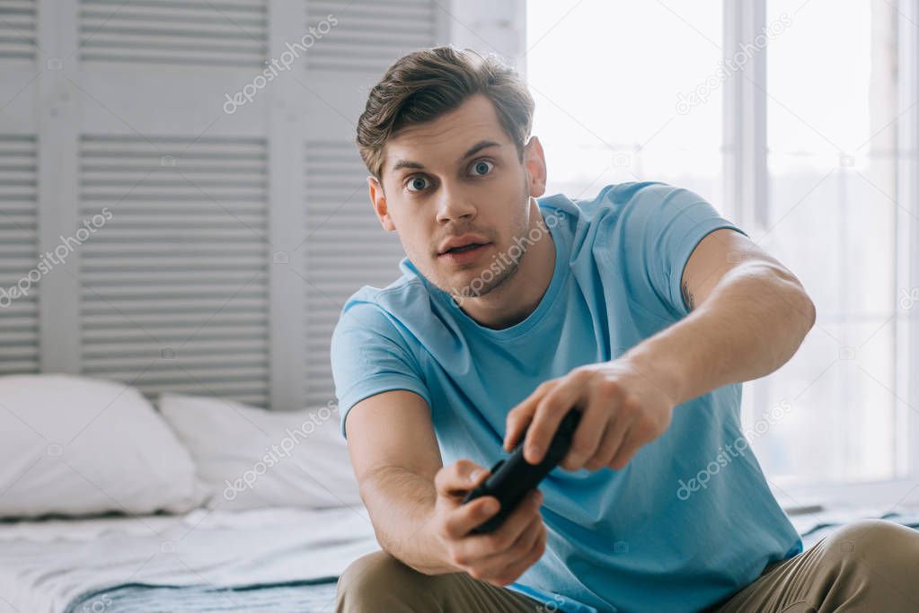 Surprised young man with joystick playing video game while sitting on bed