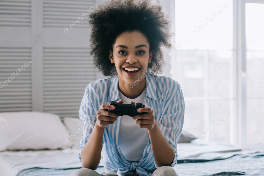 African american woman with joystick playing video game while sitting on bed
