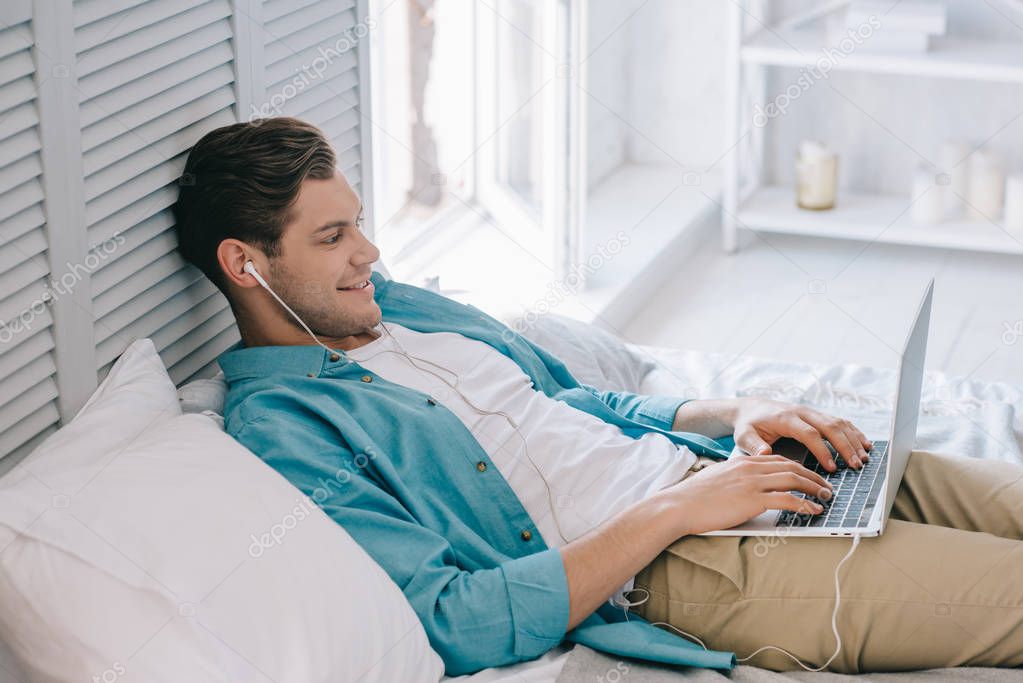 Smiling man in earbuds using laptop while lying on bed at home