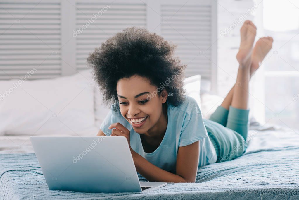 Smiling african american girl lying on bed and looking at laptop screen
