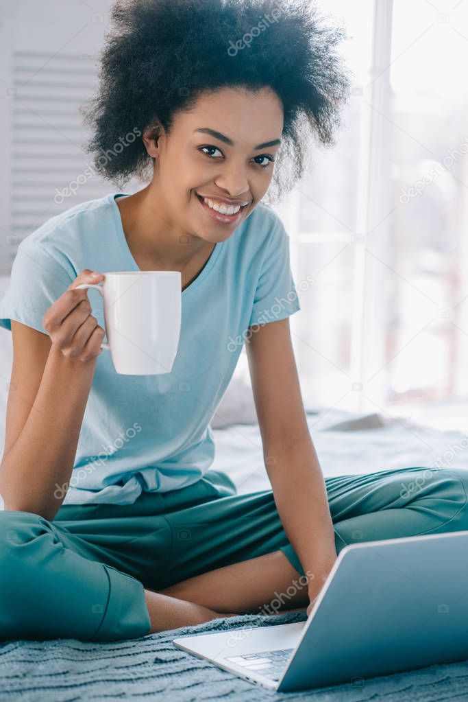 Smiling african american girl in lounge wear holding cup of coffee and using laptop in bed