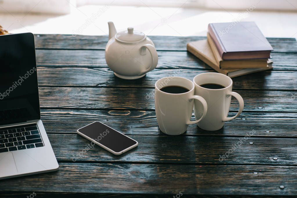 Cups and teapot on wooden table with laptop and smartphone