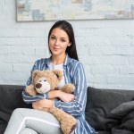 Portrét usmívající se žena s plyšovým medvědem v rukou sedí na gauči doma