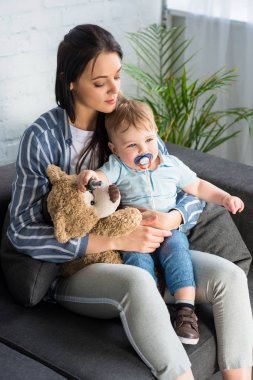 genç anne ve bebek emziği ve evde koltukta oturan bir oyuncak ayı ile