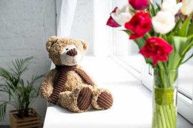 selective focus of teddy bear and bouquet of tulips in vase o window sill at home clipart
