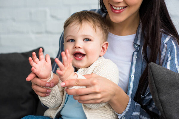 partial view of smiling mother playing with little baby at home