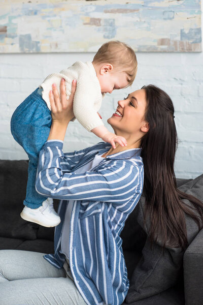 side view of happy mother with baby in hands sitting on sofa at home