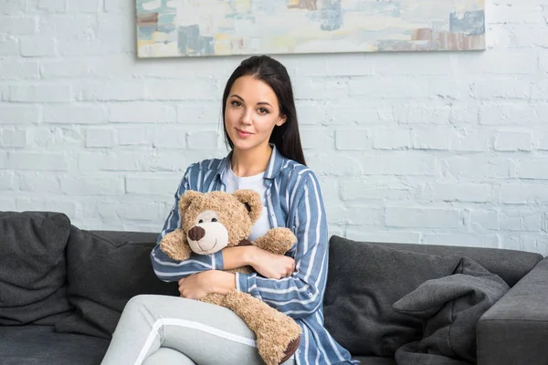 portrait of smiling woman with teddy bear in hands sitting on sofa at home