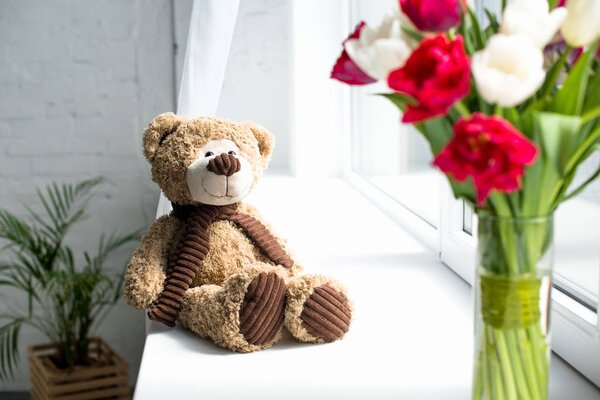 selective focus of teddy bear and bouquet of tulips in vase o window sill at home