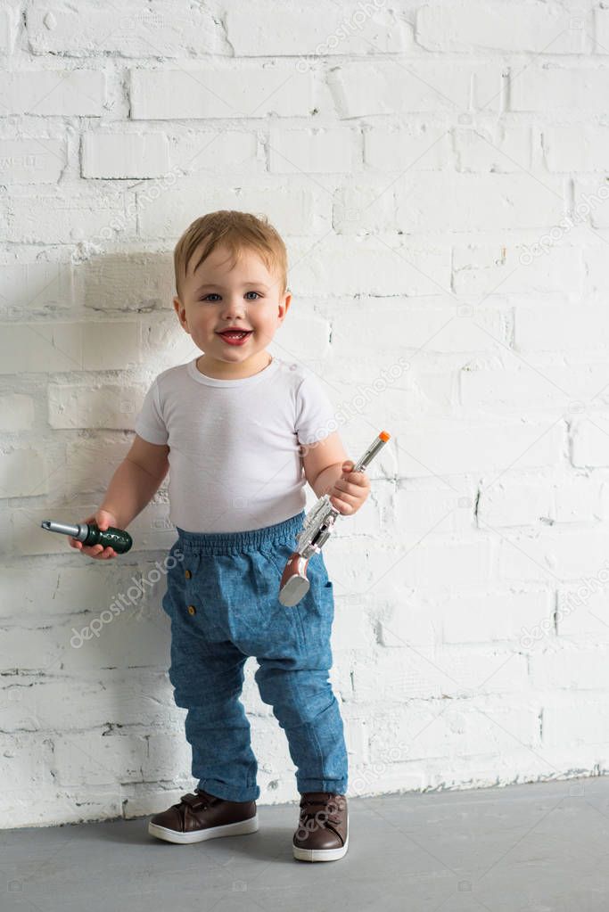 happy little baby boy with toys standing at white brick wall