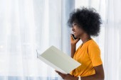 Seitenansicht eines afrikanisch-amerikanischen Freelancers mit Folder, der zu Hause auf dem Smartphone spricht