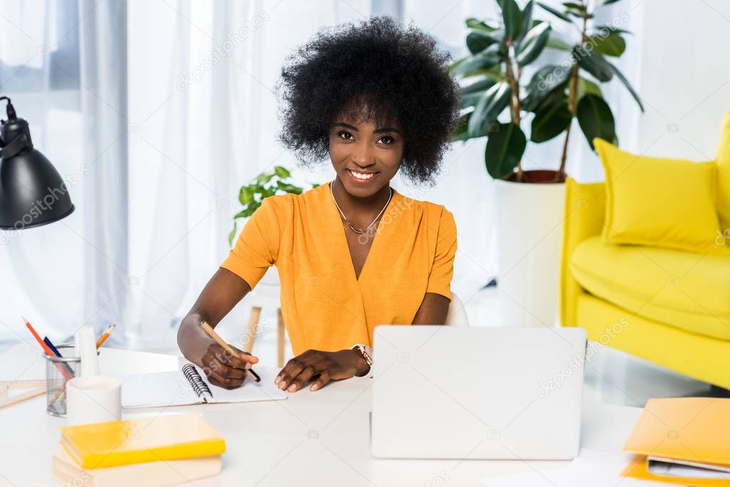 portrait of smiling african american freelancer at workplace with laptop and notebook at home
