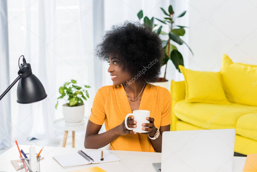 portrait of smiling african american woman with cup of coffee in hands at workplace at home
