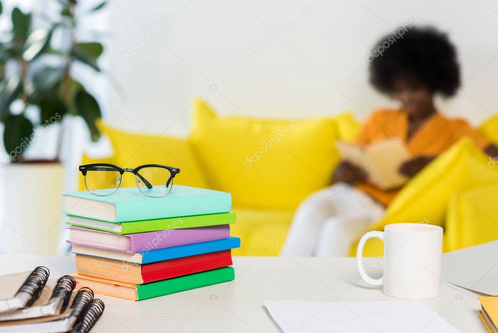 selective focus of pile of books, eyeglasses, cup of coffee at workplace and african american freelancer on sofa on background at home