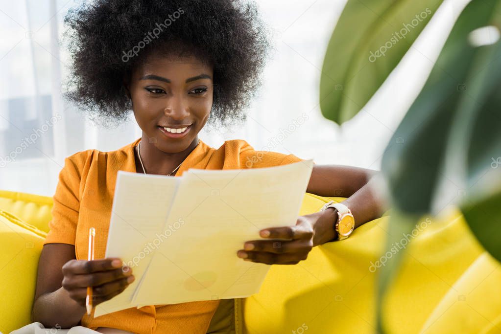 portrait of smiling african american woman with papers remote working on sofa at home