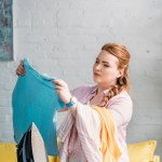 Beautiful woman looking at shirt near ironing board at home
