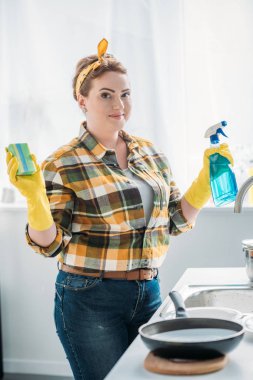 beautiful woman holding washing sponge and spray bottle while cleaning at kitchen clipart