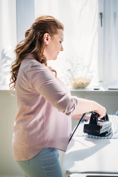 side view of beautiful woman ironing shirt with iron at home