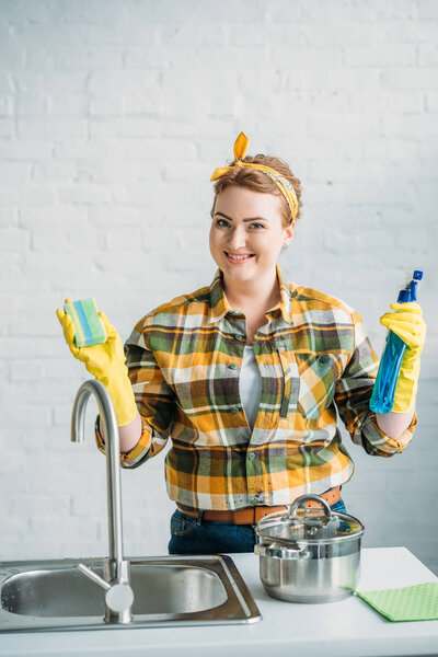 smiling woman holding washing sponge and spray bottle while cleaning at kitchen