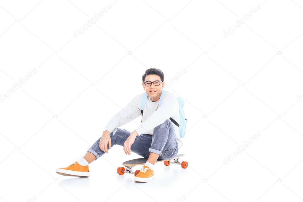 asian teenager with backpack sitting on skateboard isolated on white