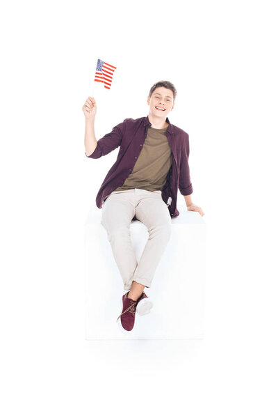 teenage student boy sitting on white cube and looking at camera with usa flag isolated on white