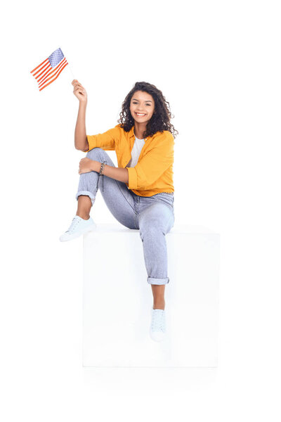 teenage african american student girl sitting on white cube and looking at camera with usa flag isolated on white