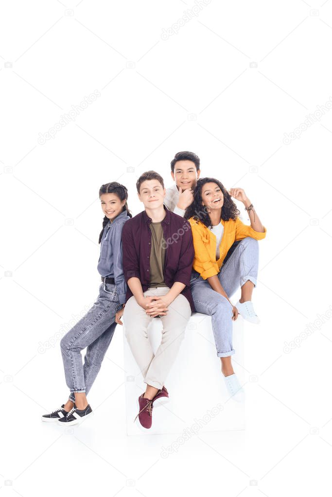 group of happy multiethnic students sitting on white cube isolated on white