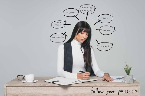 attractive brunette businesswoman writing something at table isolated on gray with speech bubbles and follow your passion inscription