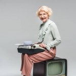 Smiling senior woman using typewriter while sitting on vintage tv on grey