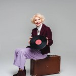 Happy elegant senior kvinna med vinyl skiva sitter på vintage resväska på grå bakgrund