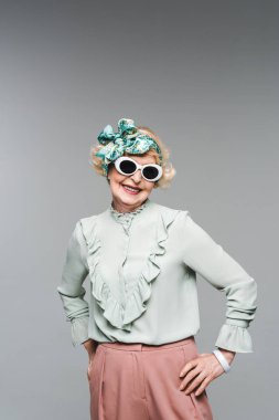 happy senior woman in stylish headband and sunglasses isolated on grey clipart
