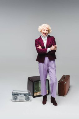 çapraz kolları vintage tv, boombox ve bavulu Grey önünde ayakta olan güzel kadın kıdemli