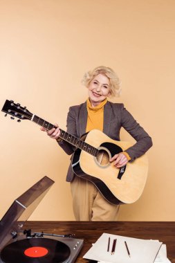 gülümseyen kıdemli kadın vinil plak çalar tabloyla yakınındaki akustik gitar çalmaya 