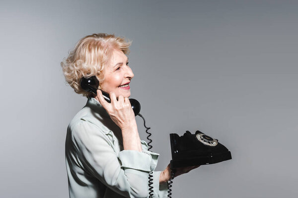 вид сбоку улыбающейся пожилой женщины, разговаривающей по винтажному телефону, изолированному на сером фоне

