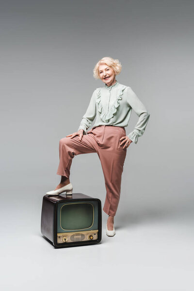 beautiful senior woman in stylish clothes with feet on vintage tv on grey
