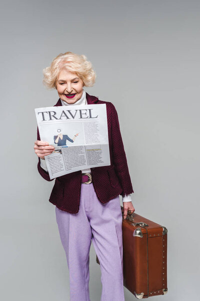 beautiful senior woman holding suitcase and reading travel newspaper isolated on grey