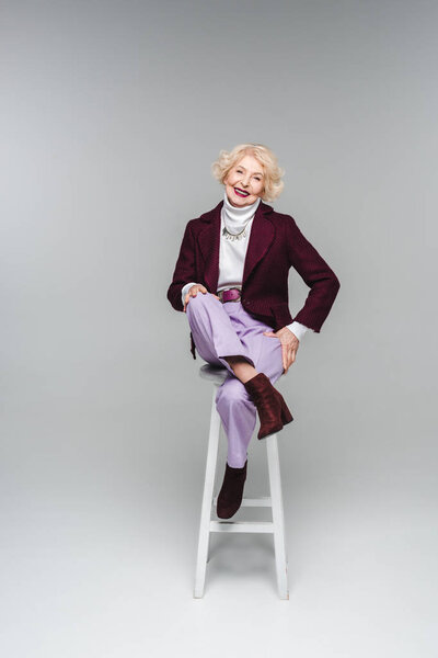 счастливая пожилая женщина в стильной одежде, сидящая на стуле на сером
