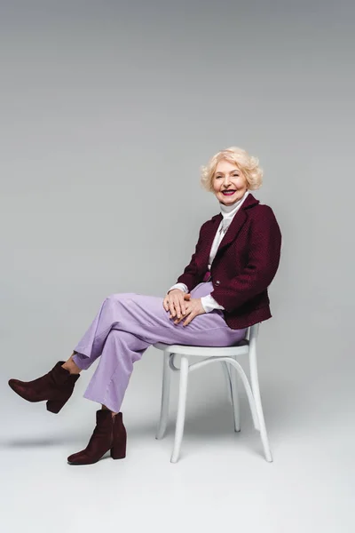 Lächelnde Seniorin Sitzt Auf Stuhl Und Blickt Kamera Auf Grau — Stockfoto
