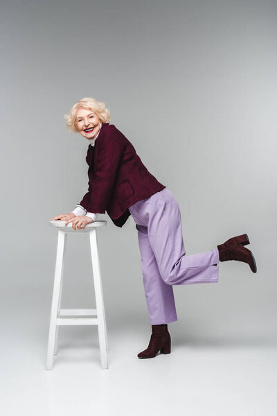 beautiful senior woman in stylish vintage clothes leaning on chair on grey