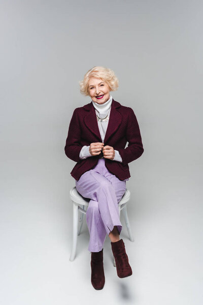 beautiful senior woman in stylish jacket sitting on chair and looking at camera on grey