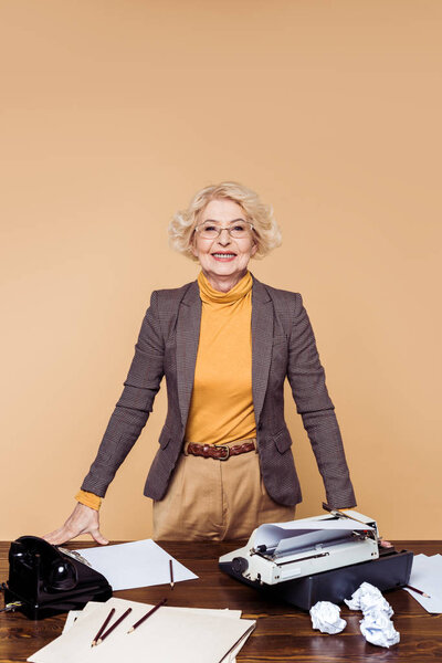 улыбающаяся пожилая женщина в очках, стоящая за столом с бумагами, печатной машинкой и ротационным телефоном
