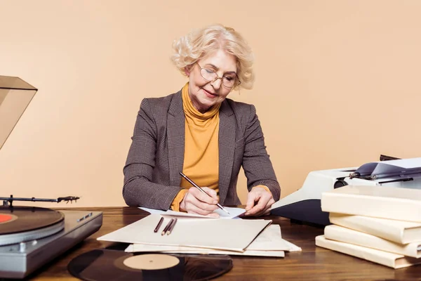 senior woman in eyeglasses writing in paper at table with vinyl disc, record player and typewriter
