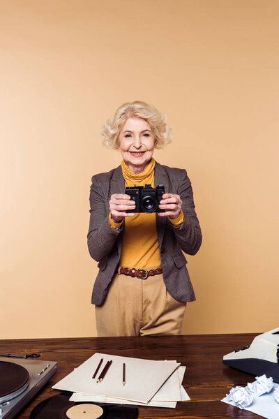 smiling stylish senior woman with film camera isolated on beige background