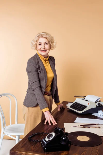 stylish senior woman standing at table with rotary phone, vinyl plate and typewriter