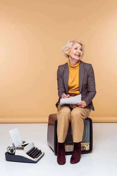 smiling stylish senior woman writing on paper sitting on vintage tv near typewriter