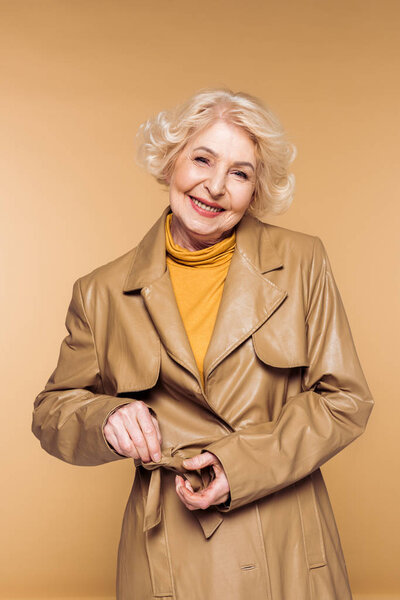smiling fashionable senior woman tying belt of leather trench coat isolated on beige background 